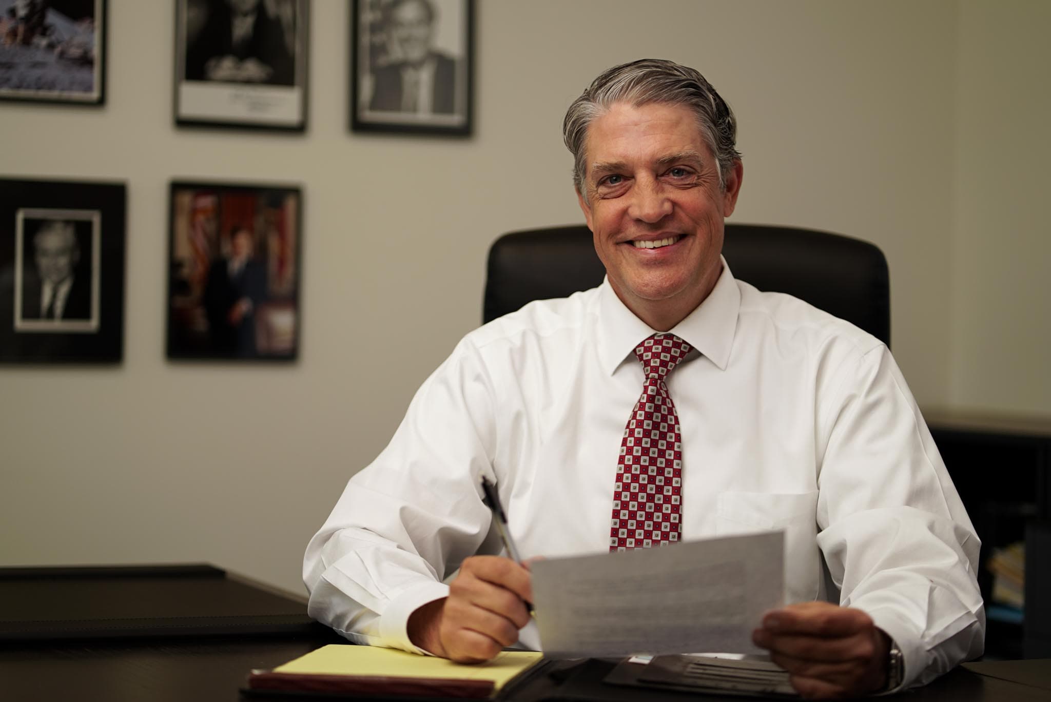 A senior Atlas attorney smiling at his desk, pen in hand, beneath a wall of portraits.