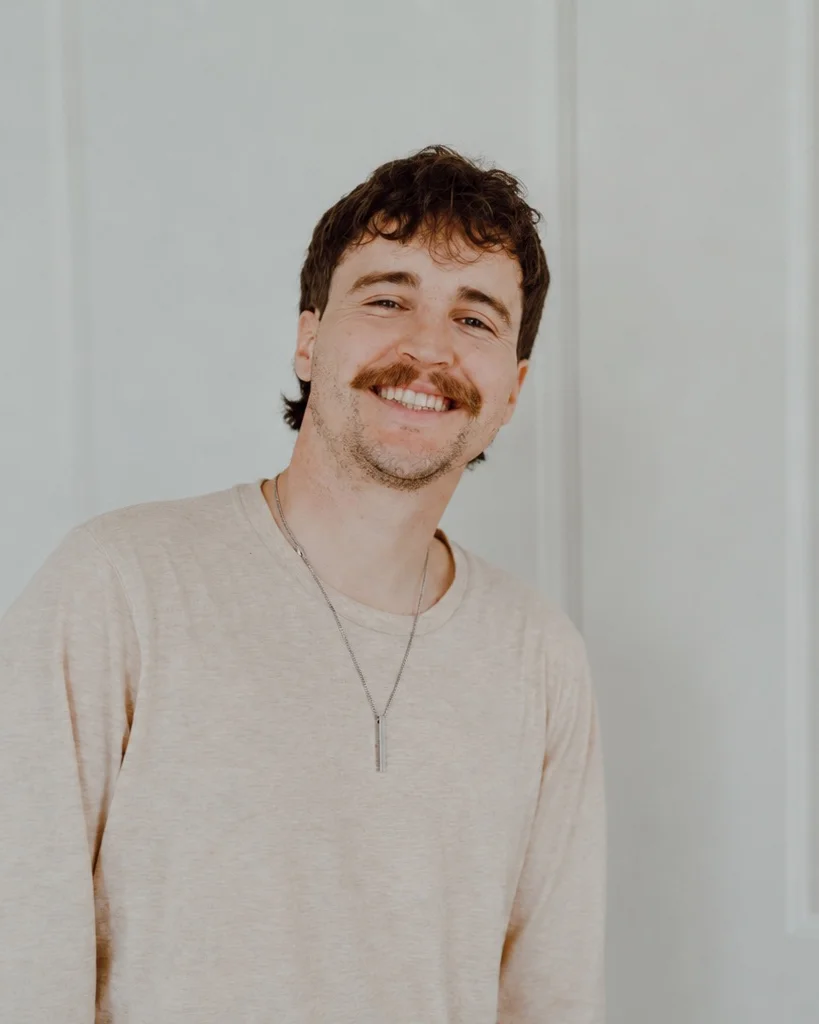 Bailey Mullens smiling in a brown jacket against a light neutral background.